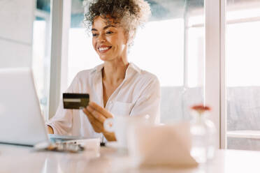 Carefree mature woman smiling cheerfully while shopping online at home. Happy mature woman using a credit card and a laptop to make an online purchase while sitting alone at a table. - JLPSF04831