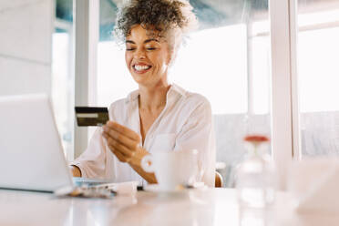 Happy mature woman smiling cheerfully while shopping online at home. Mature woman using a credit card and a laptop to make an online purchase while sitting alone at a table. - JLPSF04832