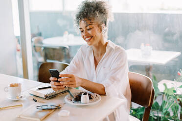 Mature businesswoman talking on a video call in a cafe. Mature businesswoman smiling while speaking with her colleagues on a smartphone. Businesswoman sitting alone at a cafe table. - JLPSF04836