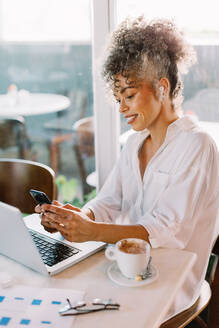 Geschäftsfrau, die ein Smartphone benutzt, während sie in einem Café arbeitet. Reife Geschäftsfrau, die fröhlich lächelt, während sie allein an einem Cafétisch sitzt. Glückliche Geschäftsfrau, die eine Textnachricht liest. - JLPSF04837