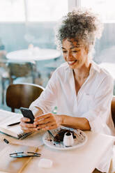 Cheerful mature businesswoman taking a video call in a cafe. Mature businesswoman smiling while speaking with her colleagues on a smartphone. Businesswoman sitting alone at a cafe table. - JLPSF04838