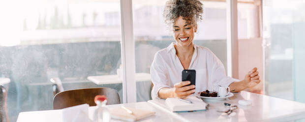 Erfolgreiche reife Geschäftsfrau, die ein Smartphone in einem Café benutzt. Glückliche Geschäftsfrau, die fröhlich lächelt, während sie allein an einem Cafétisch sitzt. Geschäftsfrau, die eine Textnachricht von ihrem Telefon liest. - JLPSF04840