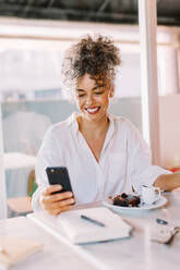 Moderne reife Geschäftsfrau, die ein Smartphone in einem Café benutzt. Glückliche Geschäftsfrau, die fröhlich lächelt, während sie allein an einem Kaffeetisch sitzt. Geschäftsfrau, die eine Textnachricht von ihrem Telefon liest. - JLPSF04841