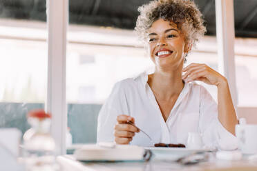 Glückliche reife Frau, die mit einem Lächeln auf dem Gesicht wegschaut. Reife Frau, die Kuchen isst, während sie an einem Cafétisch sitzt. Fröhliche Frau, die alleine in einem Café zu Mittag isst. - JLPSF04844