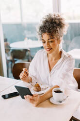 Reife Geschäftsfrau, die einen Videoanruf während des Mittagessens annimmt. Glückliche reife Geschäftsfrau, die ein digitales Tablet hält, während sie allein an einem Cafétisch sitzt. Geschäftsfrau, die einen Cupcake und Kaffee in einem Café isst. - JLPSF04850