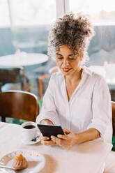 Geschäftsfrau, die ein digitales Tablet benutzt, während sie in einem Café Kaffee trinkt. Reife Geschäftsfrau, die im Internet surft, während sie in der Mittagspause allein an einem Cafétisch sitzt. - JLPSF04851