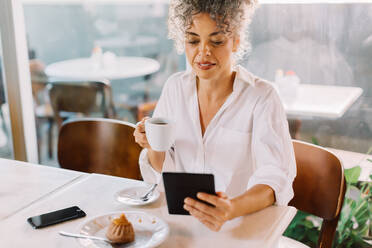 Moderne Geschäftsfrau, die ein digitales Tablet benutzt, während sie in einem Café Kaffee trinkt. Reife Geschäftsfrau, die im Internet surft, während sie in der Mittagspause allein an einem Cafétisch sitzt. - JLPSF04852