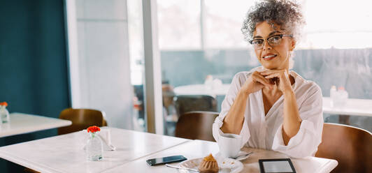 Fröhliche Geschäftsfrau, die mit einem leichten Lächeln in die Kamera schaut, während sie allein in einem Café sitzt. Reife Geschäftsfrau mit Brille beim Mittagessen in einem Café. - JLPSF04859