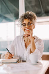 Smiling businesswoman looking at the camera while working in a cafe. Mature businesswoman making notes and planning her week in her work journal while sitting alone at a cafe table. - JLPSF04860