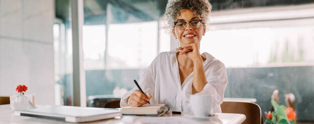 Successful businesswoman smiling at the camera while working in a cafe. Mature businesswoman making notes and planning her week in her work journal while sitting alone at a cafe table. - JLPSF04861