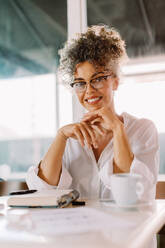 Confident businesswoman smiling at the camera while sitting alone in a cafe. Successful mature businesswoman sitting with her work journal on the table in a coffee shop. - JLPSF04862