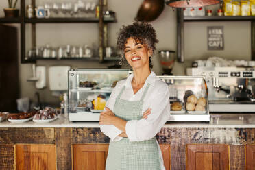 Happy mature business owner smiling at the camera while standing with her arms crossed. Successful businesswoman feeling confident while wearing an apron in front of her coffee shop during the day. - JLPSF04864