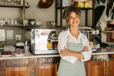 Cheerful business owner smiling while standing with her arms crossed. Successful mature businesswoman feeling confident while wearing an apron in front of her coffee shop during the day. - JLPSF04866