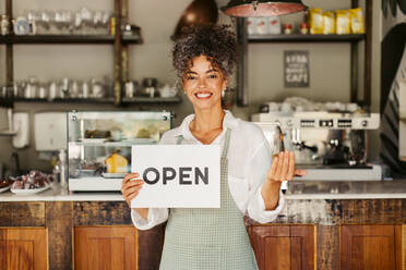 Mature business owner holding an open sign in front of her newly opened cafe. Happy mature businesswoman smiling at the camera while wearing an apron at her new coffee shop. - JLPSF04867