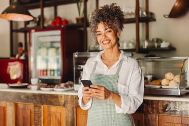 Erfolgreiche Café-Besitzerin, die in ihrem Café ein Smartphone benutzt. Reife Geschäftsfrau, die in die Kamera lächelt, während sie vor dem Tresen steht. Geschäftsfrau, die eine Textnachricht sendet. - JLPSF04868