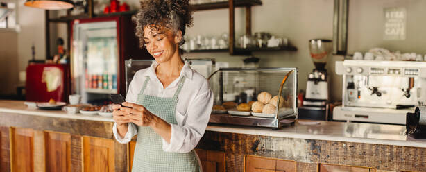 Cafe owner using a smartphone in her cafe. Mature business owner smiling cheerfully while standing in front of the counter. Successful businesswoman communicating with her customers online. - JLPSF04870