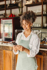 Reife Café-Besitzerin, die ein Smartphone in ihrem Café benutzt. Reife Geschäftsinhaberin, die fröhlich lächelt, während sie vor dem Tresen steht. Erfolgreiche Geschäftsfrau, die eine Textnachricht liest. - JLPSF04871