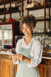 Geschäftsinhaberin, die in ihrem Café ein Smartphone benutzt. Reife Café-Besitzerin, die fröhlich lächelt, während sie vor der Theke steht. Erfolgreiche Unternehmerin, die mit ihren Kunden online kommuniziert. - JLPSF04872