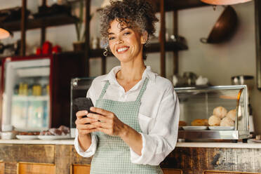 Kleinunternehmerin, die in ihrem Café ein Smartphone benutzt. Reife Geschäftsfrau, die in die Kamera lächelt, während sie vor dem Tresen steht. Erfolgreiche Unternehmerin, die online mit ihren Kunden kommuniziert. - JLPSF04873