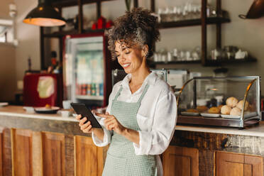 Glückliche Geschäftsinhaberin, die ein digitales Tablet in ihrem Café benutzt. Reife Café-Besitzerin, die fröhlich lächelt, während sie vor der Theke steht. Erfolgreiche Unternehmerin, die ihr Geschäft online betreibt. - JLPSF04874