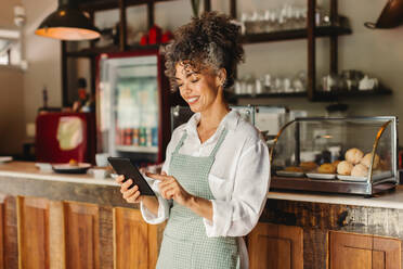 Happy business owner using a digital tablet in her cafe. Mature female cafe owner smiling cheerfully while standing in front of the counter. Successful entrepreneur running her business online. - JLPSF04874