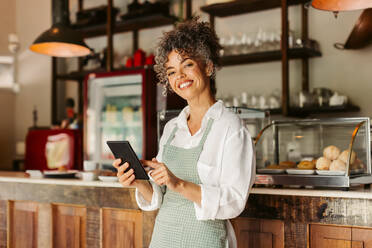 Entrepreneur holding a digital tablet in her cafe. Mature female cafe owner smiling at the camera while standing in front of the counter. Successful businesswoman running her business online. - JLPSF04875