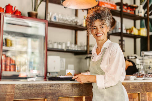 Freundliche Café-Besitzerin, die ein Tablett mit Essen in ihrem Café hält. Reife Barista-Frau, die fröhlich lächelt, während sie Kunden in einem Café bedient. Reife Geschäftsinhaberin, die allein arbeitet. - JLPSF04877