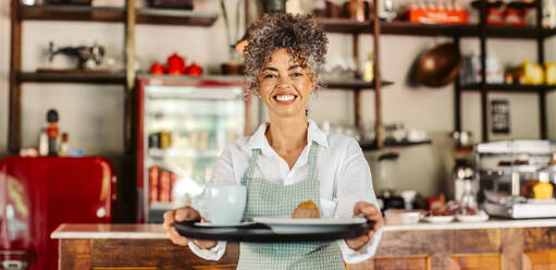 Glückliche reife Café-Besitzerin, die einem Kunden ihre Bestellung gibt. Fröhlich lächelnde Barista-Frau, die einen Kunden in ihrem Café bedient. Reife Frau, die allein in einem Café arbeitet. - JLPSF04879