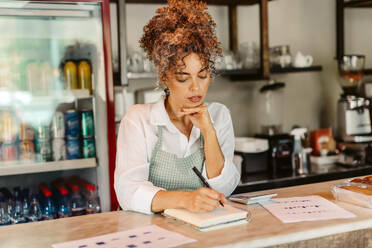 Unternehmerin, die in ihrem Café ihre monatlichen Ausgaben berechnet. Ältere Geschäftsfrau, die ihr Budget in einem Notizbuch plant. Geschäftsfrau, die sich Notizen macht, während sie allein hinter dem Tresen steht. - JLPSF04881