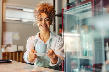 Unbekümmerte reife Barista, die einem Kunden eine Tasse Kaffee gibt. Weibliche Café-Besitzerin lächelt fröhlich, während sie einen Kunden in ihrem Café bedient. Reife Frau, die allein in einem Café arbeitet. - JLPSF04886