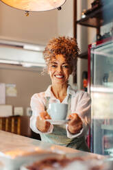 Selbstbewusste reife Barista, die einem Kunden eine Tasse Kaffee gibt. Weibliche Café-Besitzerin lächelt fröhlich, während sie einen Kunden in ihrem Café bedient. Reife Frau, die allein in einem Café arbeitet. - JLPSF04887