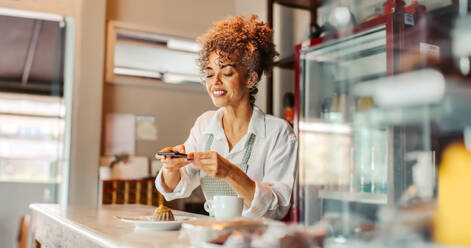 Eine Geschäftsfrau fotografiert ihr Essen mit einem Smartphone in ihrem Café. Eine reife Geschäftsfrau bewirbt ihre Speisekarte in ihrem Blog. Eine Geschäftsfrau vermarktet ihr Geschäft online. - JLPSF04893