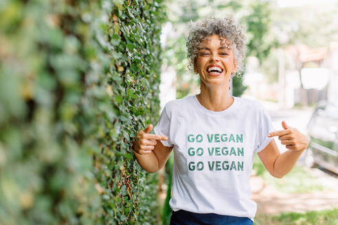 Selbstbewusste reife Veganer-Aktivistin, die fröhlich lächelt, während sie alleine im Freien steht. Fröhliche reife Frau, die für Veganismus eintritt, während sie ein T-Shirt mit der Aufschrift 