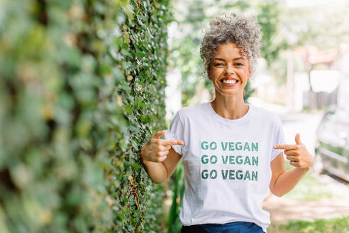 Fröhliche reife vegane Aktivistin, die fröhlich lächelt, während sie alleine im Freien steht. Fröhliche reife Frau, die für Veganismus eintritt, während sie ein T-Shirt mit der Aufschrift 