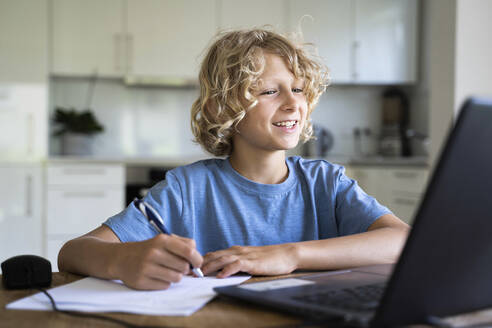 Glücklicher Junge macht Hausaufgaben und schaut zu Hause auf den Laptop - NJAF00022