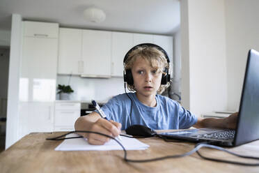 Thoughtful boy with wired headphones studying through laptop at home - NJAF00028