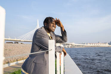 Businessman shielding eyes and leaning on railing by sea - VPIF07498