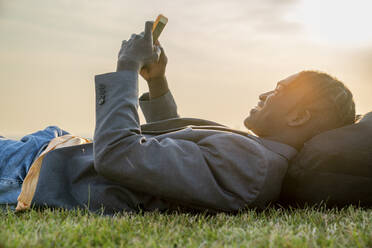 Happy young businessman using smart phone lying on grass - VPIF07523