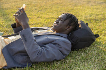 Smiling young businessman using smart phone lying on grass - VPIF07524