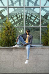 Young businessman sitting on wall in front of glass house - VPIF07531