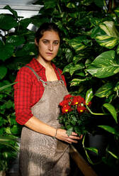 Junge Gärtnerin in kariertem rotem Hemd und grauem Overall steht inmitten von Grünpflanzen und hält rote Margaritablüten in den Händen, während sie im Gewächshaus arbeitet - ADSF39314