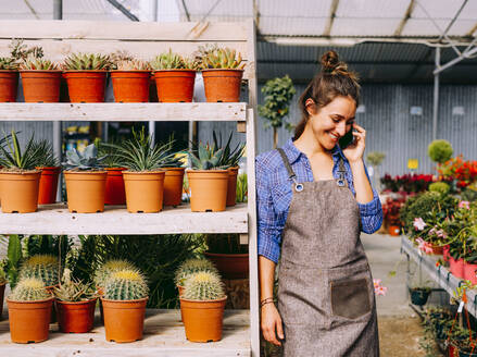 Happy lady in apron smiling and answering phone call while leaning on shelves with succulents during work in hothouse - ADSF39328