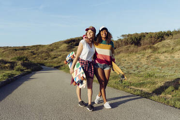 Zwei schöne multiethnische fröhliche Frauen, die Skateboards halten, während sie an einem sonnigen Tag mitten auf einer Straße laufen - ADSF39344