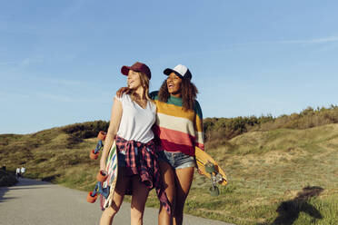 Zwei schöne multiethnische fröhliche Frauen, die Skateboards halten, während sie an einem sonnigen Tag mitten auf einer Straße laufen - ADSF39345