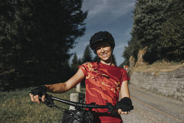 Smiling woman wearing helmet standing with bicycle at roadside - DMGF00854