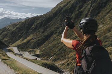 Frau mit Helm fotografiert den Gotthardpass mit dem Smartphone an einem sonnigen Tag - DMGF00865