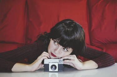 Nachdenkliche Frau, die sich vor einer roten Couch auf eine analoge Kamera stützt - SVCF00177