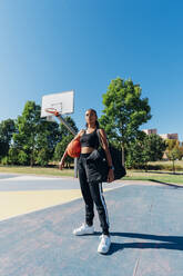 Selbstbewusste Sportlerin mit Basketball auf einem Sportplatz an einem sonnigen Tag - MEUF08173