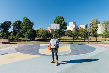 Junger Sportler mit Basketball auf einem Sportplatz an einem sonnigen Tag - MEUF08174