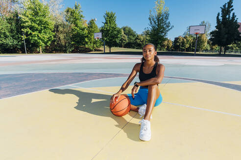 Selbstbewusste Sportlerin mit Basketball auf dem Platz sitzend - MEUF08187